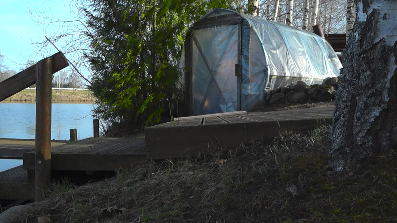 Tilt-up view of a self-made plastic sauna by the shore of lake or pond. In front of the rustic sauna wooden path and boat bridge leading into lake. Countryside retreat and unwinding by the spring lake