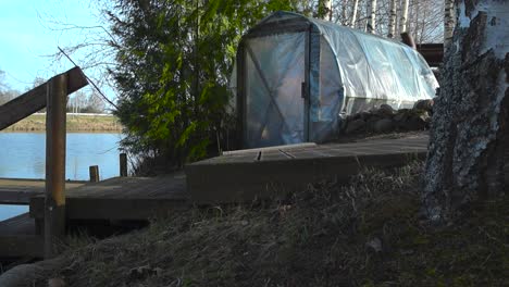 tilt-up view of a self-made plastic sauna by the shore of lake or pond. in front of the rustic sauna wooden path and boat bridge leading into lake. countryside retreat and unwinding by the spring lake