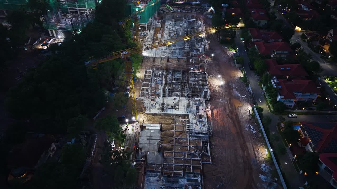 Orbiting drone shot of an illuminated construction site in Tay Ho, Hanoi, Vietnam, showing cranes, building frameworks, and a nearby residential area beside West Lake