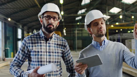 two caucasian men wearing helmets walking, talking and looking at the tablet screen in a factory