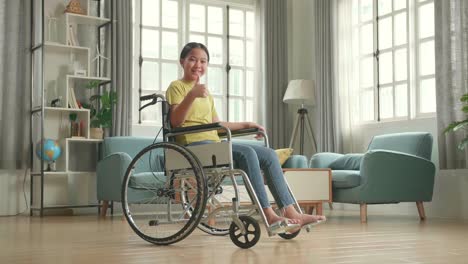 asian kid girl sitting in a wheelchair smiling and showing thumbs up in living room