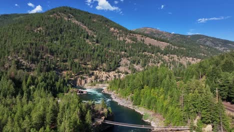 Aerial-View-Of-Kootenay-River,-Suspension-Bridge,-Forest,-And-Mountain-On-Sunny-Day-In-Libby,-Montana,-United-States