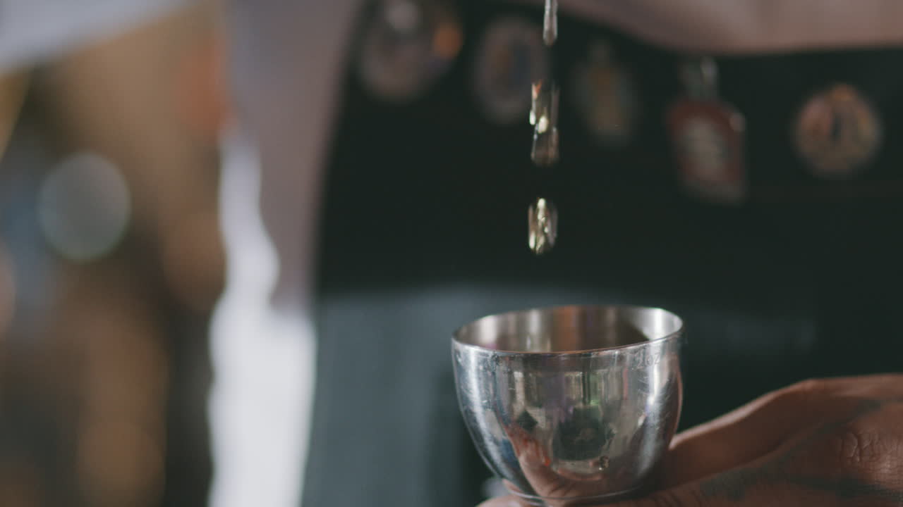 Extreme close-up showing the liquid of an alcoholic beverage being poured precisely into a metal jigger.