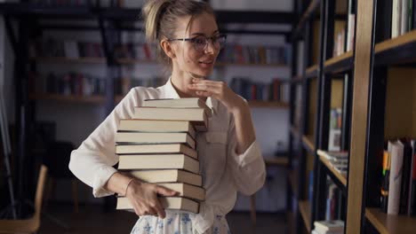 mujer positiva sosteniendo una pila de libros en la biblioteca, poniendo más en la pila