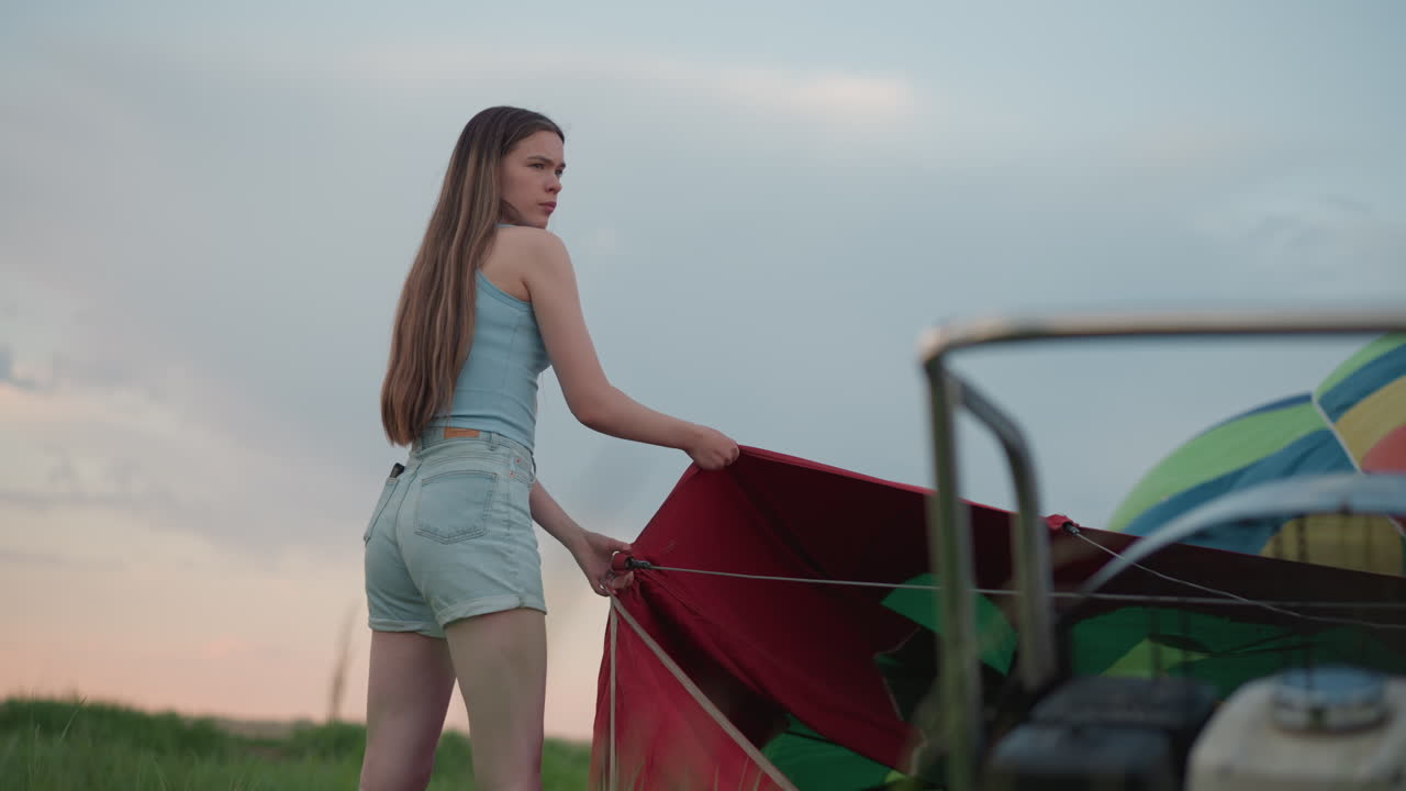 close up of rotating hot air balloon engine fan forcing wind through vibrant red envelope held by young woman in denim shorts during preflight setup in open grassy field