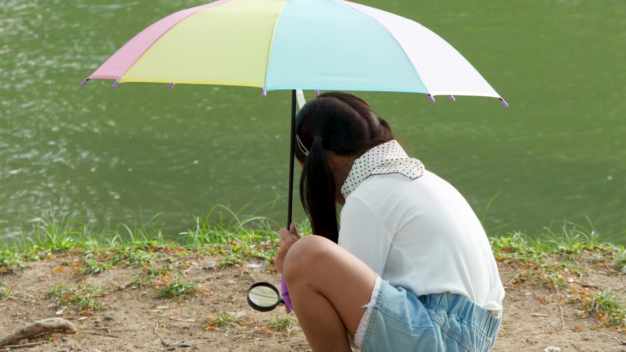 Una giovane ragazza si accovaccia vicino a un lago, usando una lente d'ingrandimento sotto un ombrello colorato