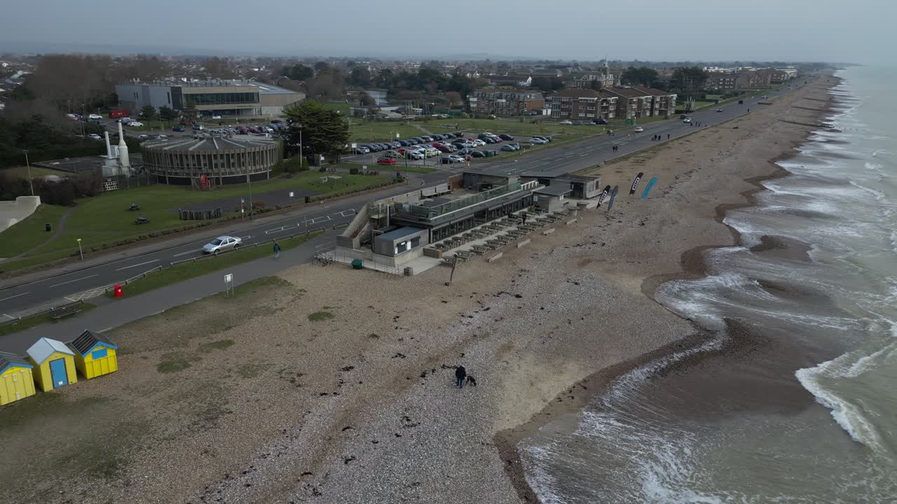 Overcast day on a pebble beach in Brighton, UK. Gentle waves wash the shore. Colorful beach huts line the beach next to a road.