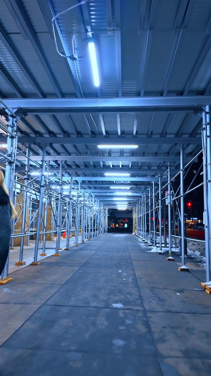 Pedoni sotto un'impalcatura sul marciapiede di notte a New York City. La vista stradale verticale mostra supporti metallici, luci aeree, pavimentazione bagnata, auto parcheggiata e neve lungo il cordolo