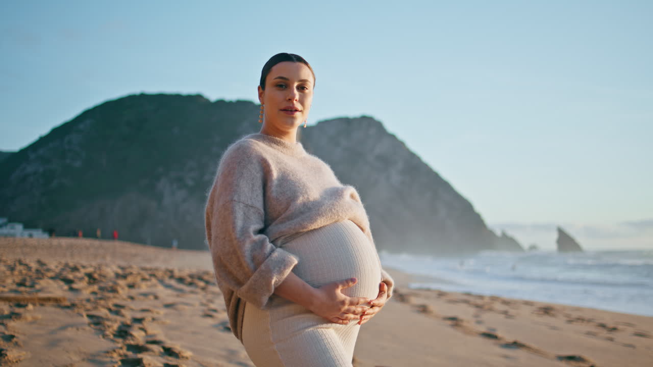 Pregnant girl posing seashore with happy smile. Expectant mother standing beach.