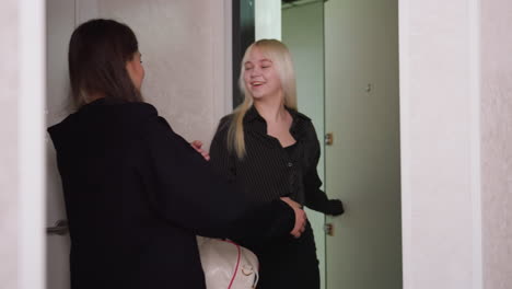 young woman in black hoodie opens door in modern hallway as her friend arrives holding handbag, warm moment follows as they greet each other with a hug, expressing joy