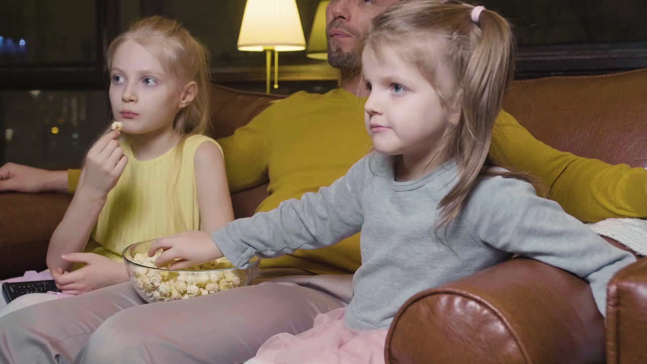 Dad And His Two Little Daughters Eating Popcorn And Watching A Movie While Sitting On Sofa At Night At Home 2