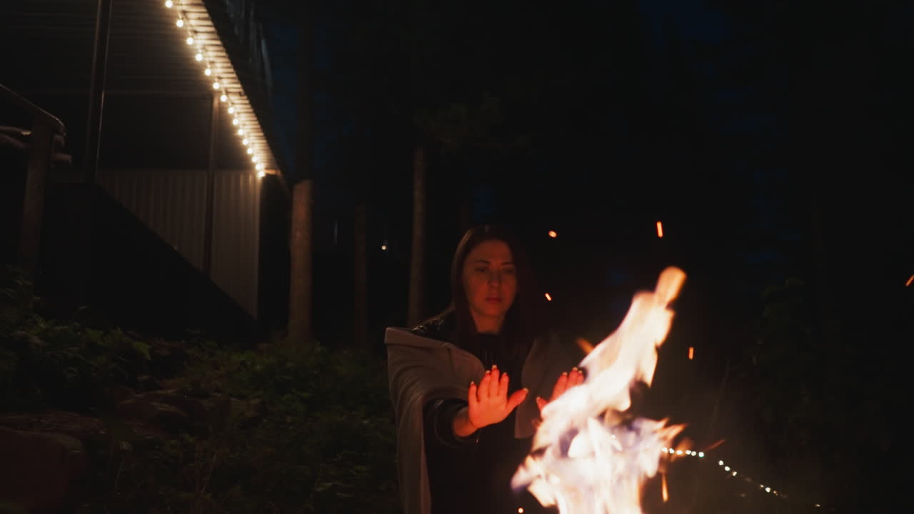 mujer calienta las manos sobre un fuego alto cerca de la casa. dama envuelta en una manta cálida trabaja para defenderse del frío de la noche frente a la noche fría. frías noches de otoño