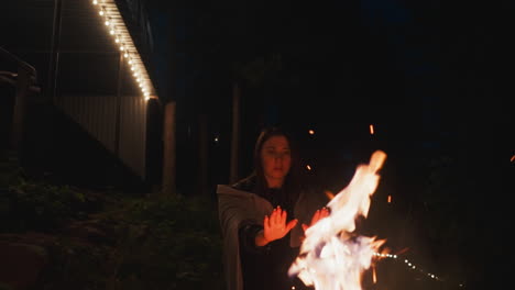 mujer calienta las manos sobre un fuego alto cerca de la casa. dama envuelta en una manta cálida trabaja para defenderse del frío de la noche frente a la noche fría. frías noches de otoño