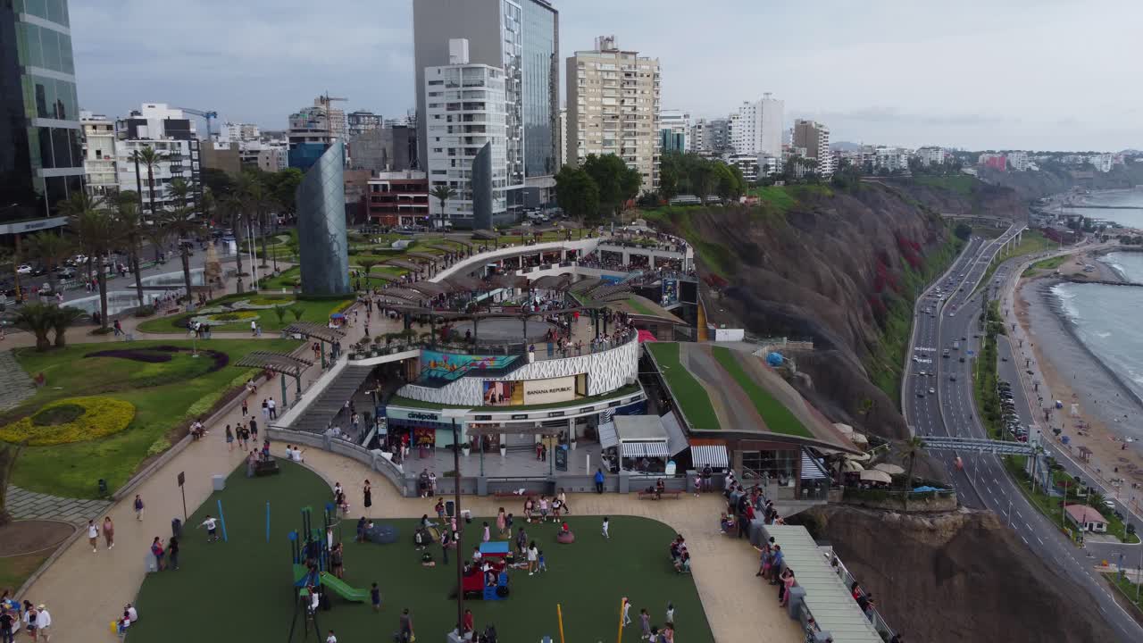 Shopping center called "Larcomar" located on the edge of a coastal cliff. Below on the right is the pacific ocean. Drone flies above and forward many people in the mall. Recorded in 4k quality.