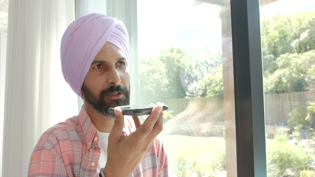 Speaking into smartphone, Indian man in turban sitting by window at home