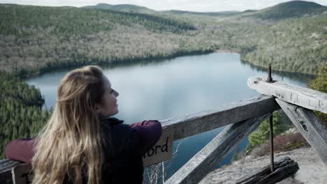 female traveler admiring beautiful scenery of calm lake and dense forest from wooden viewing platform in saint-come, quebec, canada