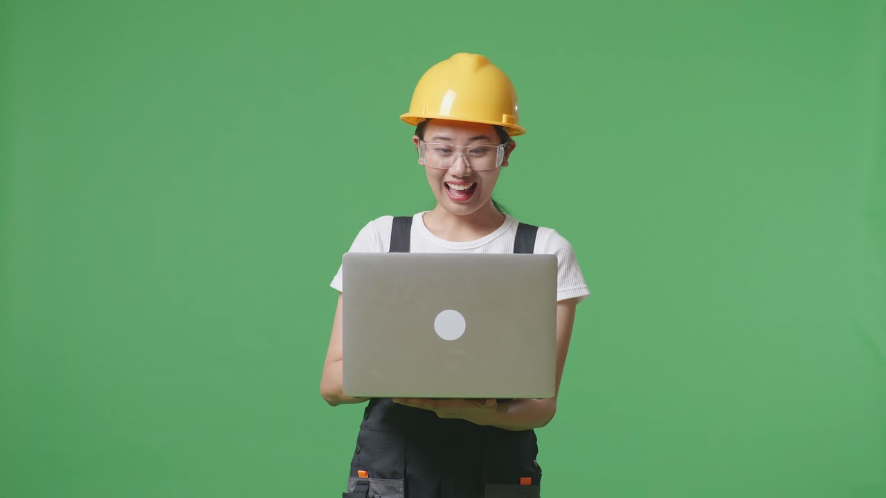 안경과 안전 헬을 입은 아시아 여성 노동자가 초록색 화면 배경 스튜디오에 서서 손에 노트북을 가리키며 미소 짓고 있습니다.