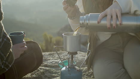 primer plano de una chica irreconocible que vierte té caliente de un termo en una taza mientras se sienta en la cima de la montaña y disfruta de un viaje de senderismo con un amigo