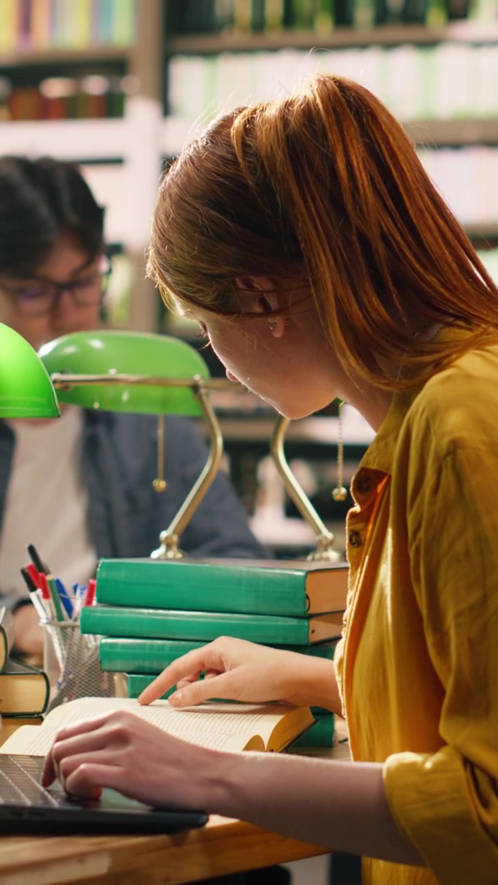 Video verticale: Studentessa dai capelli rossi che beve caffè mentre usa il laptop alla scrivania della biblioteca.
