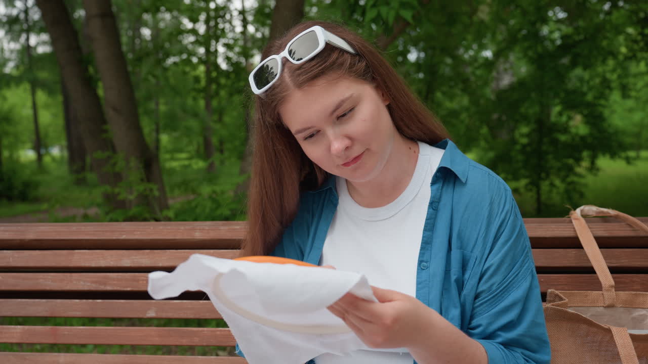 lady seated on park bench admiring embroidery work proudly while pointing at it playfully, wearing blue shirt and white outfit in green outdoor setting