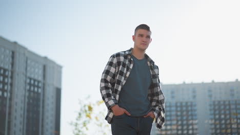 young man in checkered shirt stands with hands in pockets looking around thoughtfully under bright sky with high-rise buildings and autumn-colored tree in background on sunny urban day