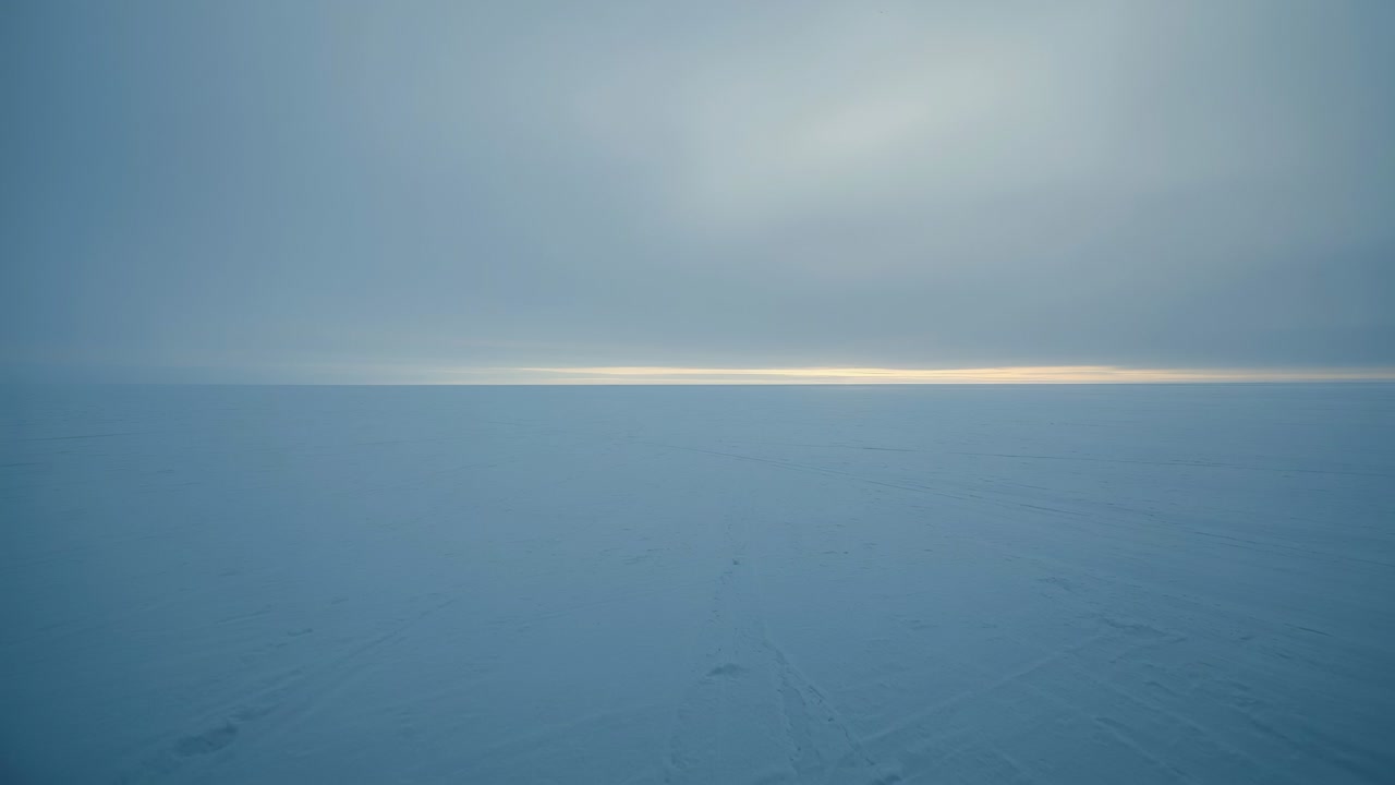 Strato di nuvole mutevole che crea una sottile fascia solare che illumina l'orizzonte su una pianura innevata e ghiacciata che mostra tracce deboli