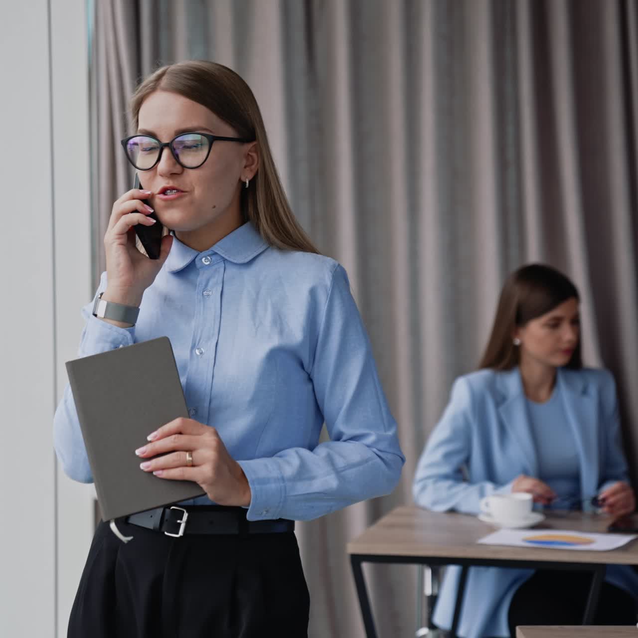 Business lady in glasses speaks on the phone standing in the office at the window. Woman is holding paper notebook in her hand. Colleagues working at backdrop