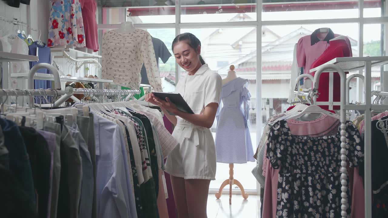 Asian Female Owner Of Clothing Store Enjoys Using Digital Tablet To Check Stock On Rails