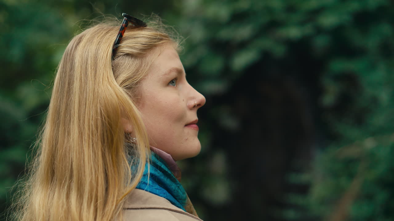 Cinematic close-up of a young woman with blonde hair looking calmly ahead, surrounded by soft green foliage in a botanical garden. Natural light, shallow depth of field and peaceful atmosphere