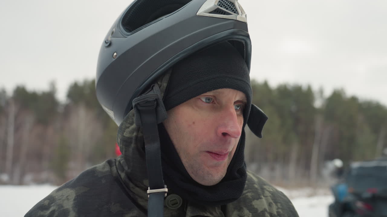 Atv rider snowy forest exhaling vapor, helmeted man adjusts chinstrap and speaks into headset, breath clouds visible in cold winter air, parked atv and pine trees in background, closeup portrait