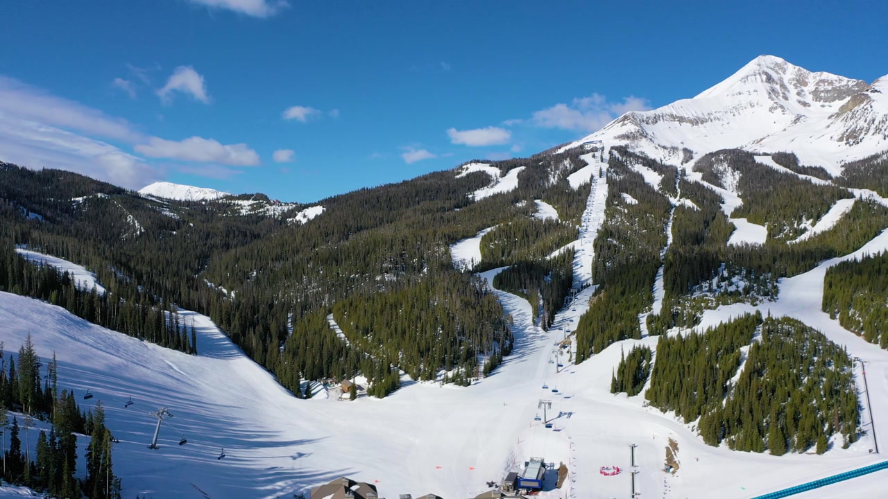 Drone shot reveals Big Sky’s chair lifts packed with skiers moving up frosty slopes as others carve down fresh powder trails framed by pine trees and icy alpine mountain peaks.