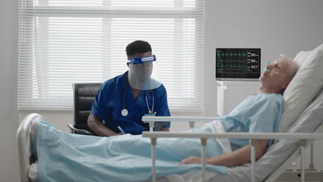 an elderly male patient lying in a hospital bed connected to an ecg machine talks to a black doctor in a protective mask