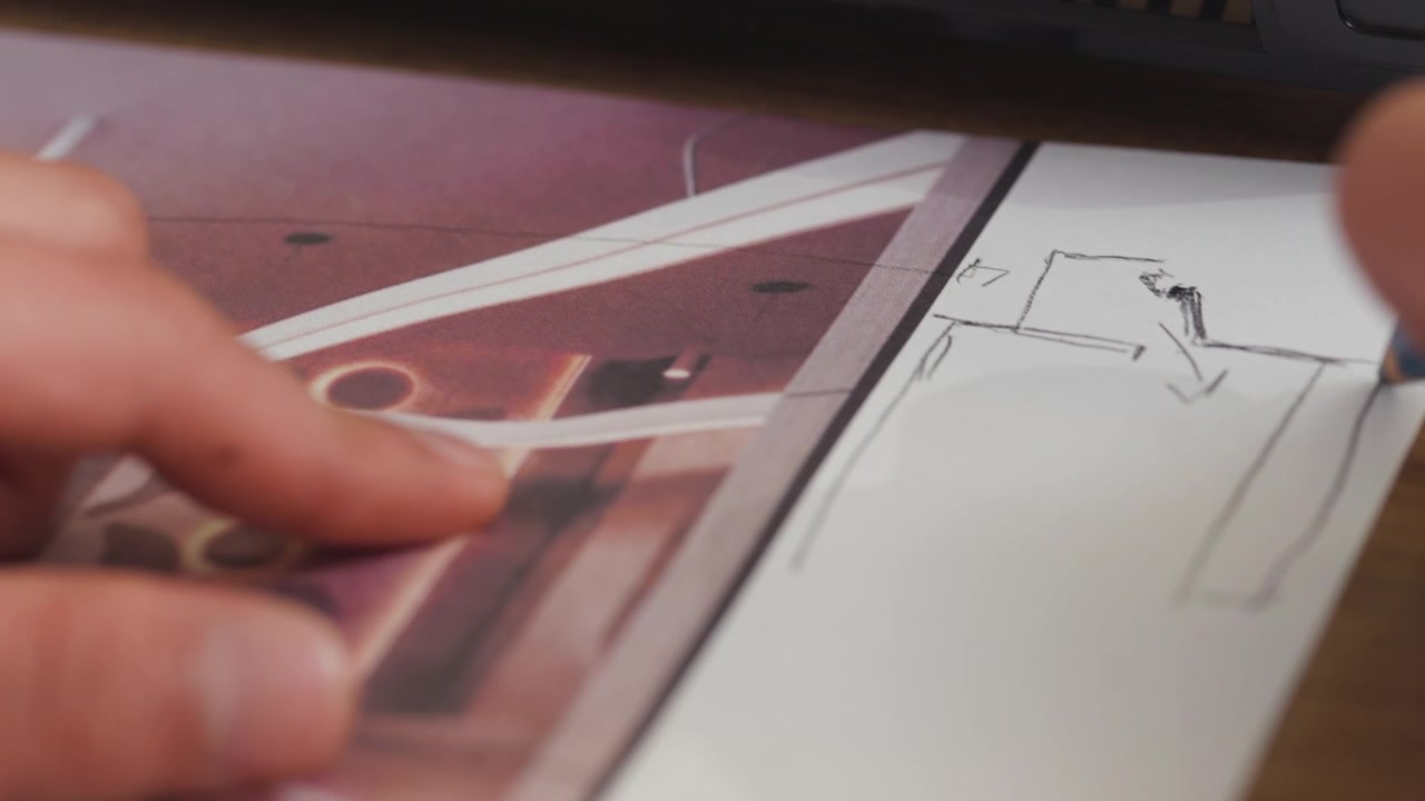 A very close-up shot showing a hand holding a pencil and tracing lines over a preliminary sketch, using a photographic print as a guide