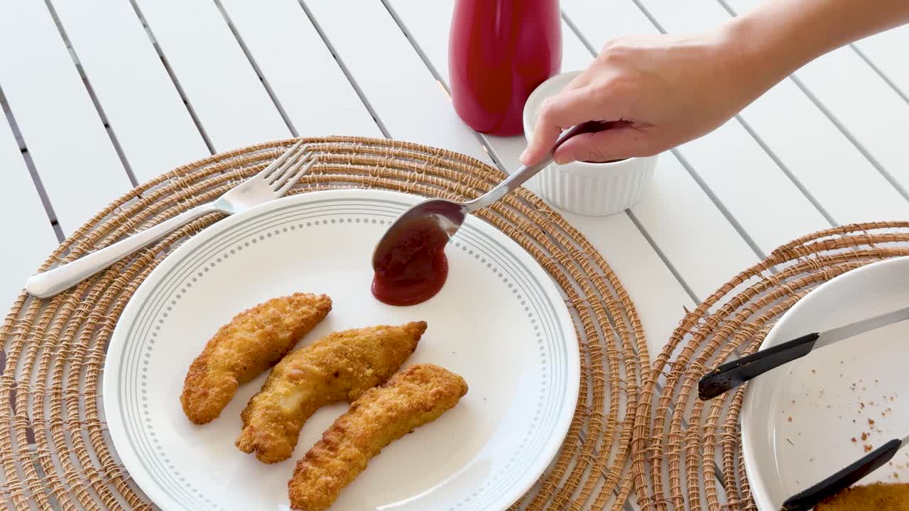 Hand scoops ketchup from ramekin and places it beside crumbed chicken tenders on a white plate, shot in bright natural lighting with overhead camera angle