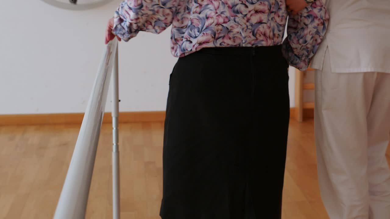 Elderly woman walking on parallel bars with the assistance of a therapist. Mobility and balance rehabilitation in a nursing home. Assisted physical therapy to regain gait and confidence