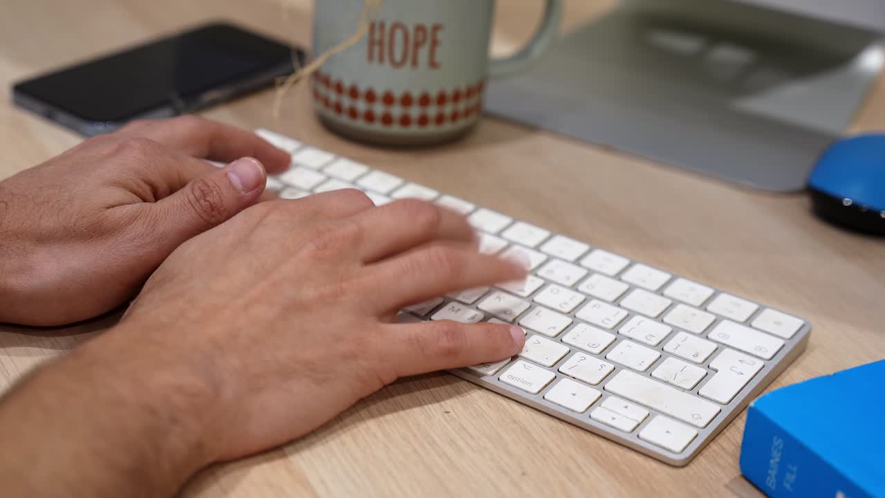 Primo piano di mani che digitano su una tastiera di computer su una scrivania di legno, con una tazza di caffè, uno smartphone e un monitor nelle vicinanze, rappresentando produttività, lavoro d'ufficio, comunicazione e attività di uno spazio di lavoro moderno.