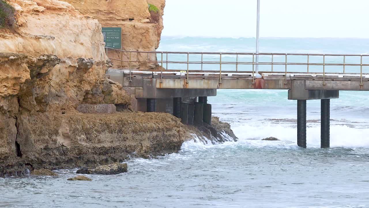 Dynamic ocean waves crash against rocky cliffs and a bridge in Port Campbell, Australia, under soft daylight