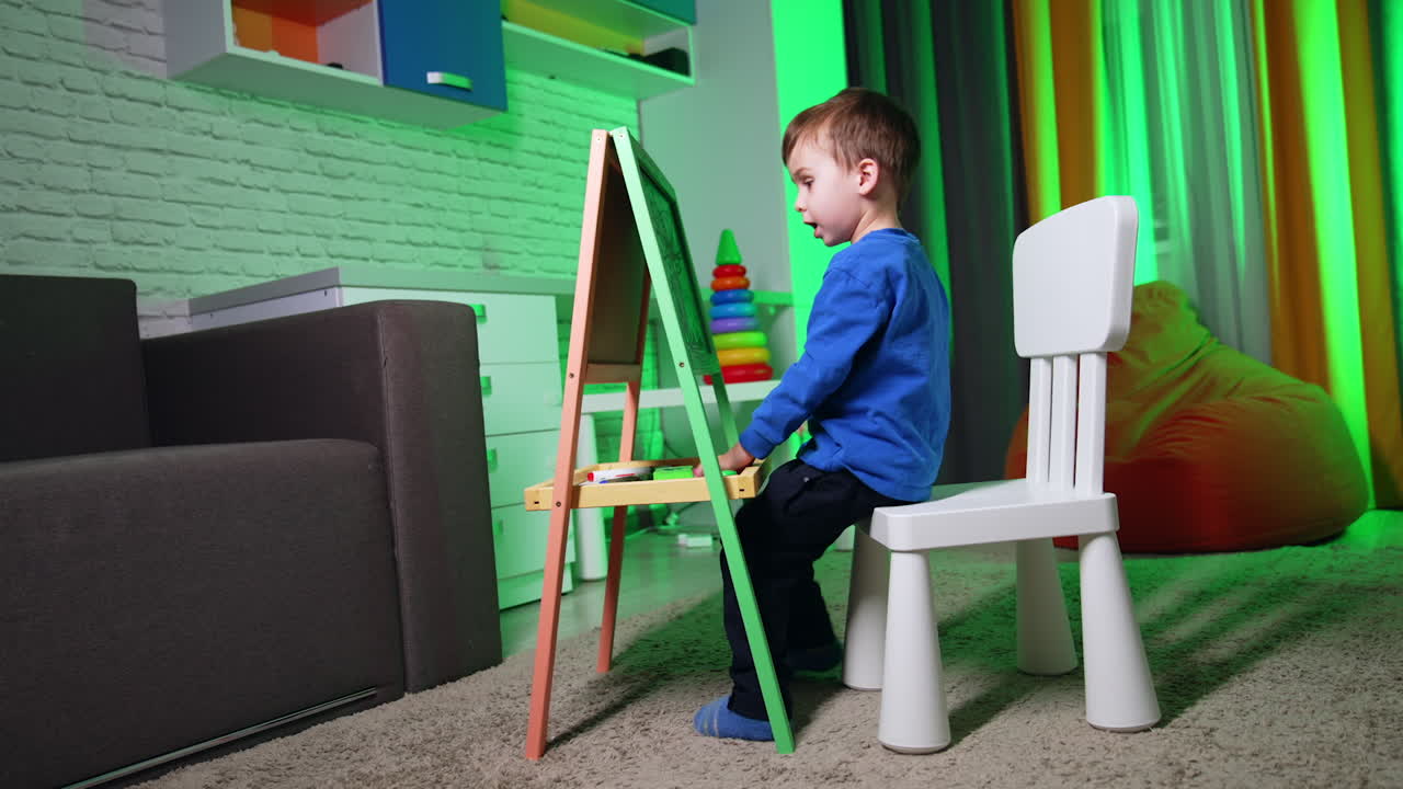 Small baby boy in blue sweater draws excitedly on the blackboard. Toddler kid doing hobby at home.
