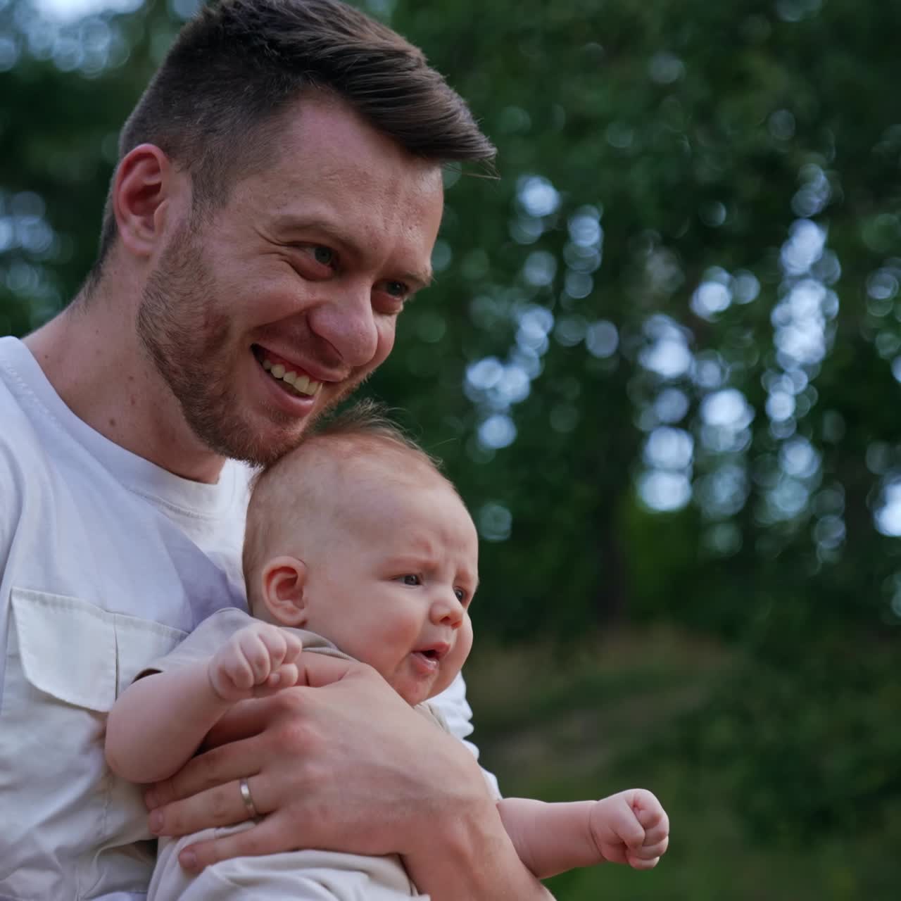 Bearded man wearing white t-shirt holding a baby. Loving dad waving his son and kissing kid's head. Blurred nature backdrop