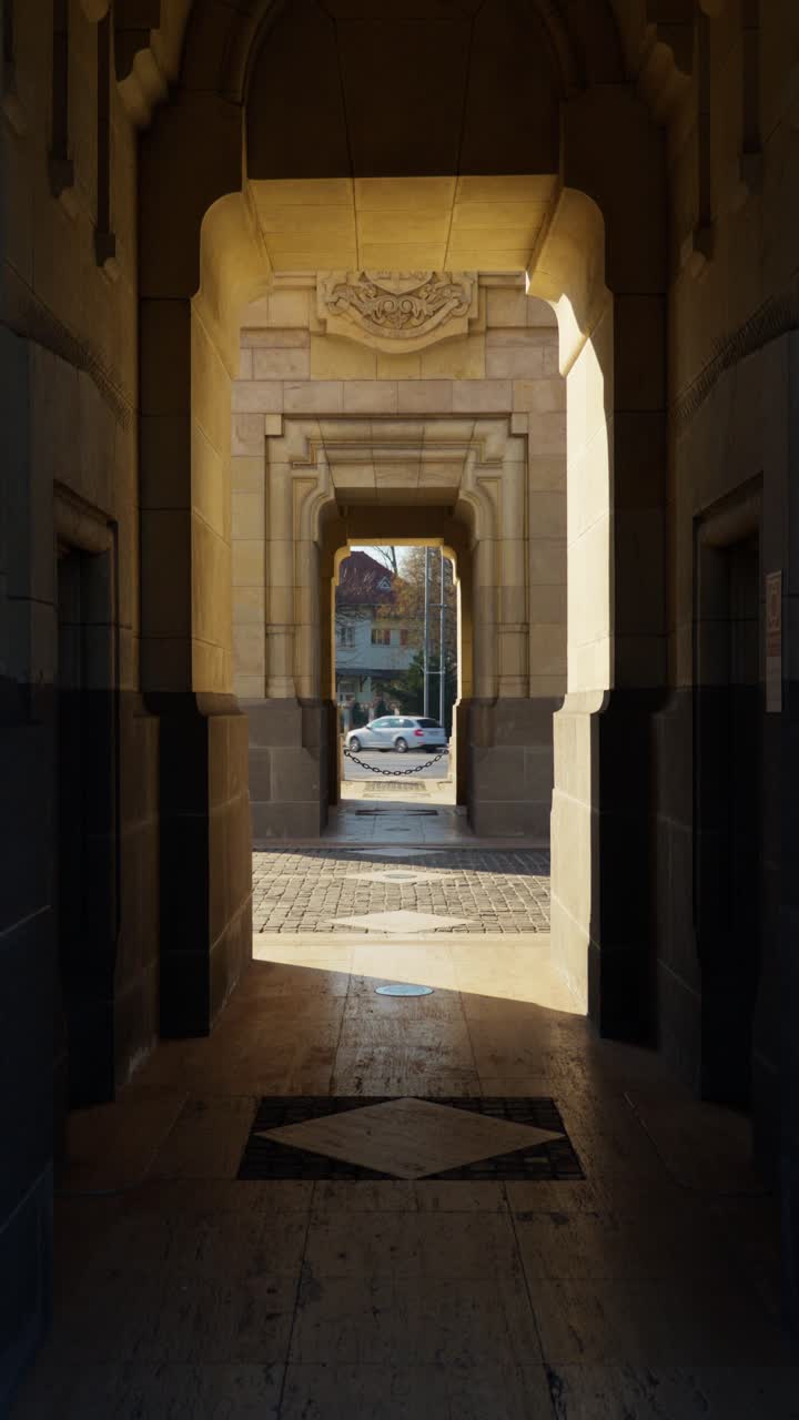 Static shot through the base of Arc de Triumf in Bucharest, sunlight casting sharp shadows on the stone passage. Framed view leads the eye to the city beyond.