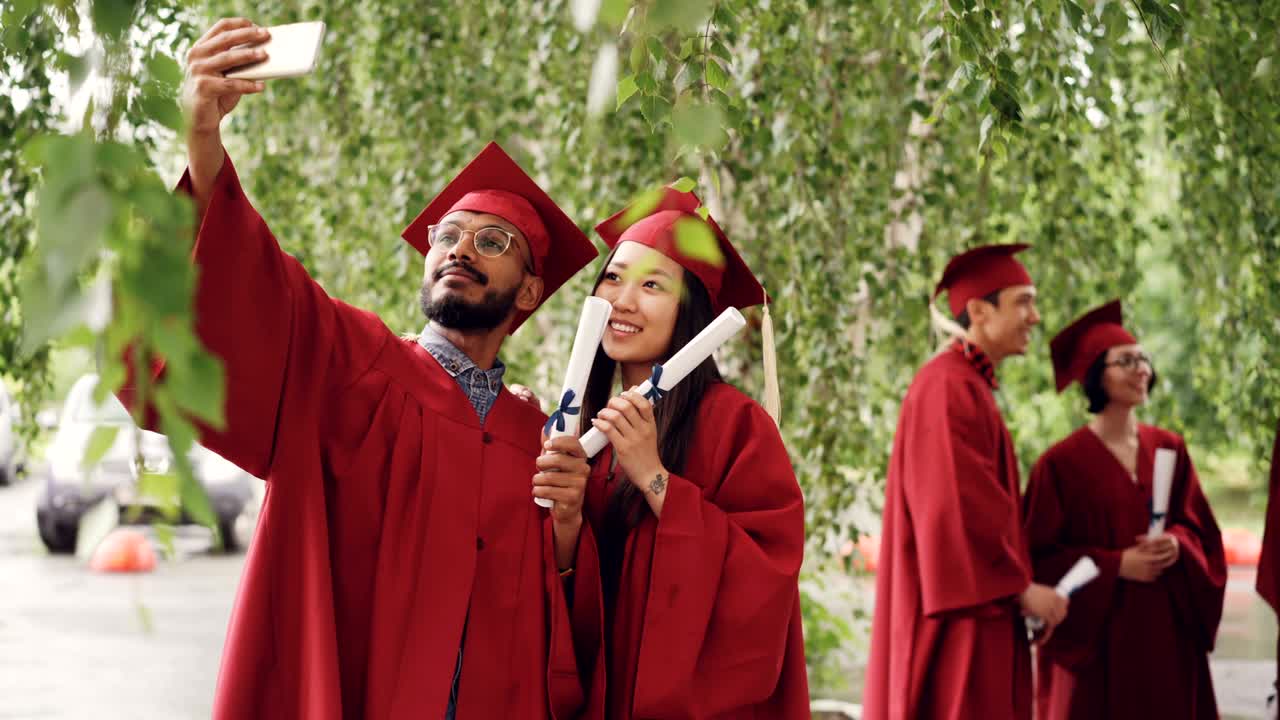 Two mixed-race students are taking selfie with graduation diplomas wearing mortarboards and gowns, guy is holding smartphone and taking picture.