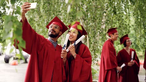 dos estudiantes de raza mixta se están tomando selfies con diplomas de graduación con cartones de mortero y vestidos, el tipo está sosteniendo un teléfono inteligente y tomando una foto.