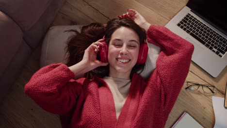 Portrait-of-a-confident-and-happy-young-brunette-girl-in-a-red-sweater-and-wireless-headphones-who-listens-to-music-during-her-break-between-work-in-a-modern-apartment-on-the-floor