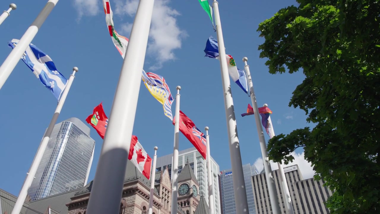 Old Toronto City Hall is gradually obscured as camera moves forward and tilts upward toward international flags waving above the square, Slow Motion