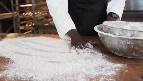 african american male baker working in bakery kitchen, spreading flour on counter in slow motion