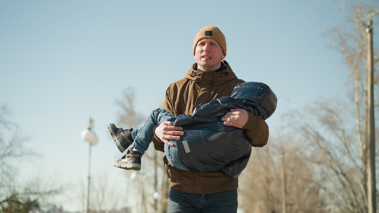 길을 는 동안 아이를 들고 있는 아버지의 클로즈업, 피곤해 보이는 아버지, 갈색 재과 베니를 입고, 아이는 검은색 부풀어있는 재을 입고 있습니다.