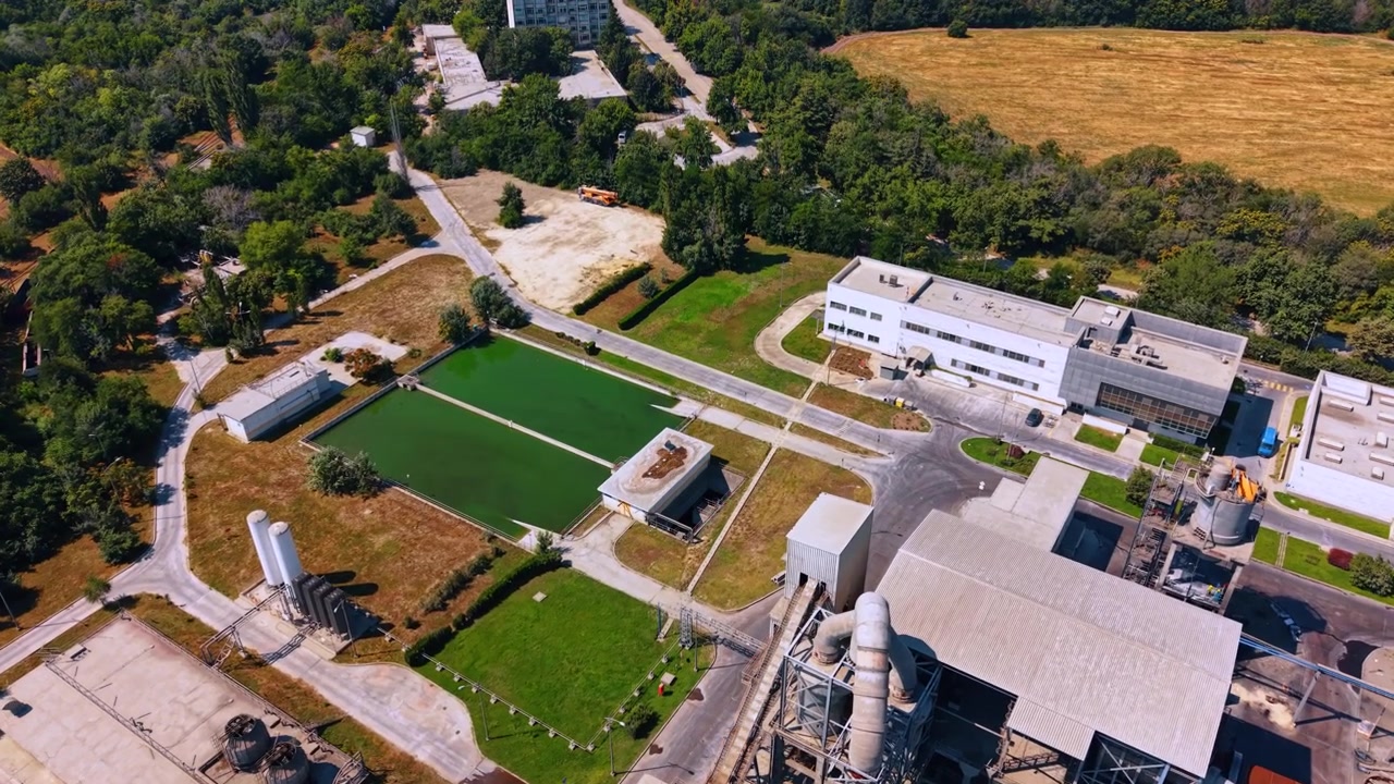 Industrivandbassiner på cementfabrikkens område. Luftfoto af to grønne damme, forsyningstanke, serviceveje, produktionsbygninger og omkringliggende træer.