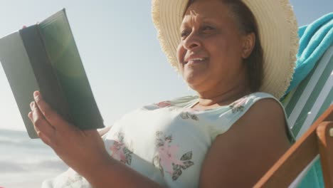 mujer afroamericana de edad avanzada sonriente acostada en una cama de sol y leyendo un libro en una playa soleada