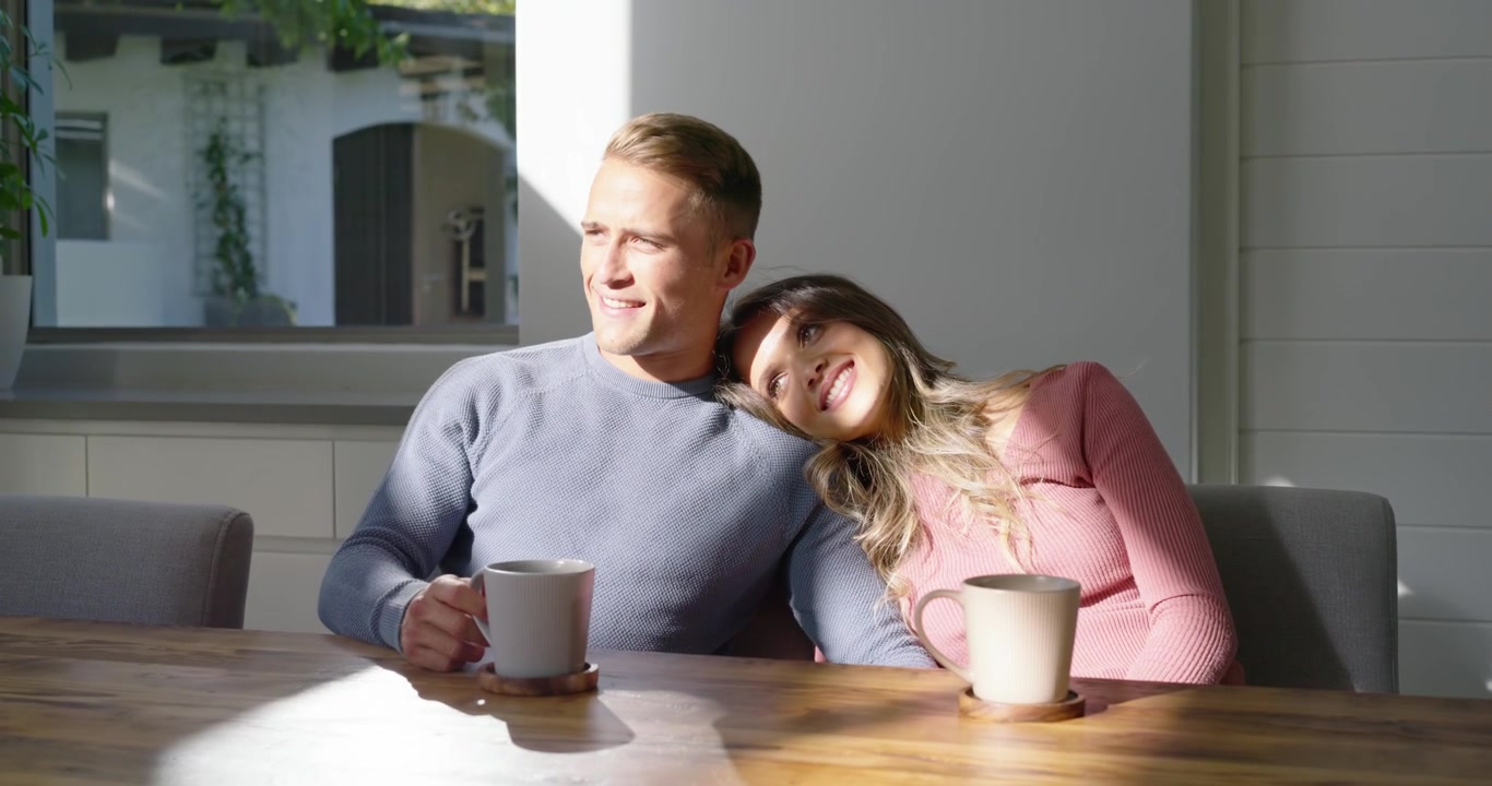 Par sidder ved bord, mand løfter krus på bordskåner, nipper, kvinde læner sig og ser morgenlyset.