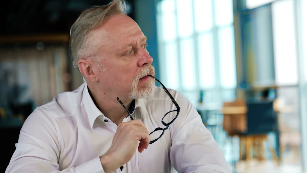 Pensive Caucasian old man sits holding his glasses. Portrait of a businessman working in office. Blurred backdrop.